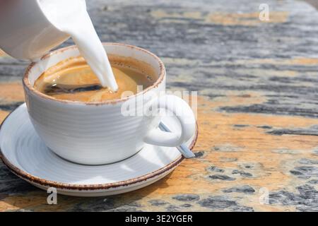 Milch in eine Tasse Americano auf rustikalem Holztisch gießen Stockfoto
