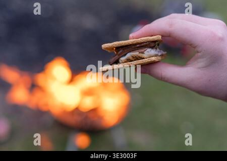 Nahaufnahme von handhaltendem Smores mit geschmolzener Schokolade und getoastetem Marshmallow in der Nähe des Lagerfeuers, gemütlichem Camping im Freien und Komfort-Food-Konzept Stockfoto