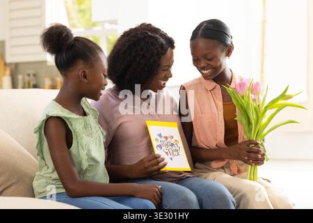 Afroamerikanische Familie, die auf einem braunen Sofa sitzt und gelbe Grußkarte liest und rosa Tulpen hält Stockfoto