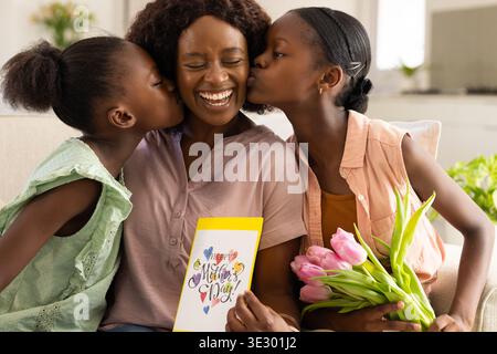 Afroamerikanische Mutter mit Jugendlichen und Mädchen, die auf dem Sofa lachen und Muttertagskarte halten, Tulpen Stockfoto
