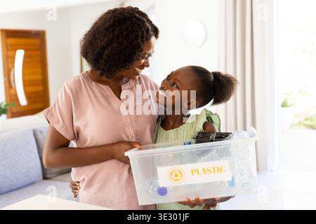 Afroamerikanische Frau und Mädchen, die zu Hause PLASTIKBEHÄLTER mit Etikett halten und Kunststoffe für das Recycling sortieren Stockfoto
