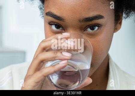 Eine Frau, die ein helles Hemd mit Kragen in der Küche trägt und klares Trinkglas hält Stockfoto