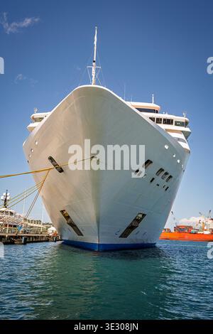 Ein großes weißes Kreuzfahrtschiff liegt in einem Hafen, dessen Bug zur Kamera gerichtet ist, mit einem Frachtschiff, das unter klarem Himmel im Hintergrund sichtbar ist Stockfoto