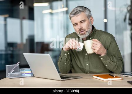 Reifer Mann sitzt an seinem Schreibtisch, hält ein Taschentuch und einen Becher, erlebt Symptome einer Erkältung oder Grippe, während er versucht, weiter zu arbeiten und hebt die Präsentation am Arbeitsplatz hervor Stockfoto