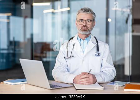 Grauhaariger männlicher Arzt mit einem Stethoskop, der an einem Schreibtisch mit Laptop und Tablet sitzt und auf die Kamera blickt und die medizinische Beratung und das Gesundheitswesen repräsentiert Stockfoto