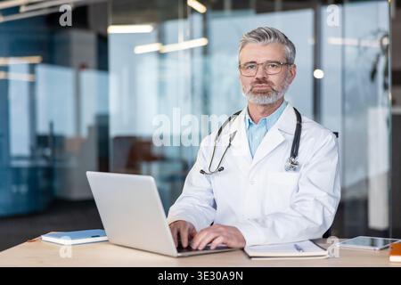 Leitender männlicher Arzt mit Brille und Stethoskop sitzt an seinem Schreibtisch, tippt auf einem Laptop, bietet professionelle medizinische Beratung und medizinische Dienstleistungen in einer klinischen Umgebung Stockfoto