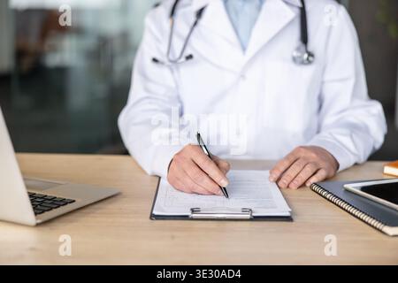 Arzt in einem weißen Mantel mit Stethoskop auf einem Klemmbrett, sitzend an einem Holzschreibtisch mit Laptop und Tablet, mit Darstellung der medizinischen Dokumentation und der Patientenversorgung Stockfoto