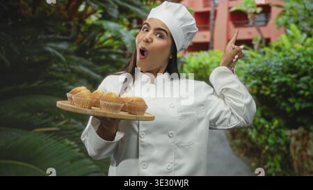 Die Chefin in weißem Mantel hält mit einer Hand ein hölzernes Muffins-Tablett und zeigt mit dem Finger auf den Weg zum Bau des Hofes; Backfreude. Stockfoto