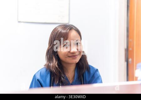 Porträt eines freundlichen medizinischen Fachmanns in blauen Peelings, die hinter einem hölzernen Schreibtisch mit verschwommenem Bokeh im Vordergrund lächeln. Gesundheits- und Medizinkonz Stockfoto