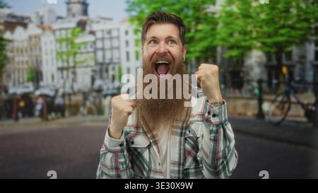 Mann mit Bart und Ohrring, der die Hände auf der Brust mit offenem Mund auf der Straße in den Gebäuden der Stadt umklammert; Erstaunen. Stockfoto