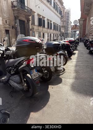 Geparkte Motorroller säumen eine sonnendurchflutete Straße in Rom, Italien, mit klassischer Architektur. Typisch italienische Nahverkehrsszene. Stockfoto