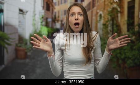 hispanische Frau mit erhobenen Händen und überraschtem Gesicht auf einer engen Straße, die von Topfpflanzen und alten Gebäuden gesäumt ist; überraschendes Verwirrungswunder. Stockfoto