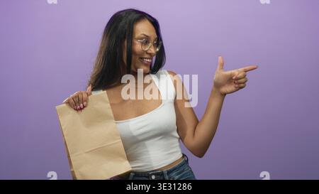 Frau mit brauner Papiertüte zeigt den Finger nach rechts und lächelt mit sichtbarer Hand vor violettem Studio-Hintergrund; verspieltes Selbstvertrauen. Stockfoto
