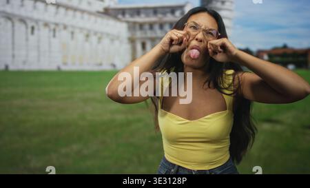 Frau zieht die Wangen und streckt die Zunge vor dem Gebäude aus, trägt gelbe Decke und Brille; verspielter Unfug. Stockfoto