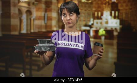 Eine Freiwillige mit kurzen Haaren hält gestapelte Folienfutterbehälter in einer Hand und eine Kaffeetasse aus Papier in der anderen in einem Kirchengebäude; müde Ser Stockfoto