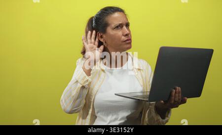 Eine brünette Frau mittleren Alters hält mit einer Hand einen Laptop und schränkt ihr Ohr mit der anderen, während sie im Studio mit gelbem Hintergrund auf den Bildschirm schwindelt Stockfoto