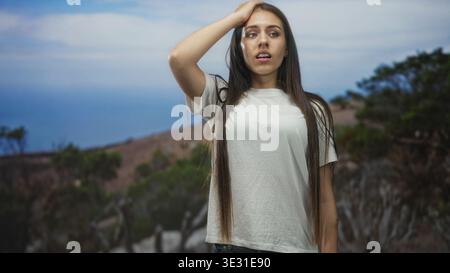 Teenager-Mädchen, das die Stirn mit Hand an der Küste berührt, in einem weißen T-Shirt und auf einer Klippe steht; ruhige Einsamkeit. Stockfoto