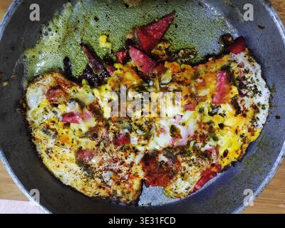Spiegeleier mit Kräutern, Wurst und rotem Pfeffer werden in einer gusseisernen Pfanne auf einem Gasherd gebraten. Stockfoto