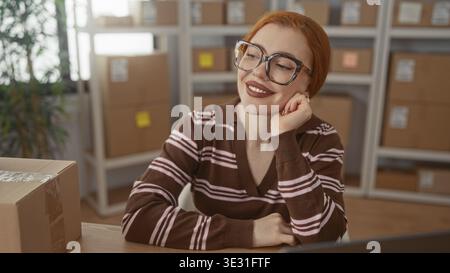 Rothaarige Frau mit Brille auf der Hand neben gestapelten Papppaketen in einem kleinen Verpackungsgebäude; unternehmerische Zufriedenheit. Stockfoto