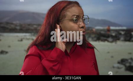 Frau, die Hand an Ohr am windigen, felsigen Strand in roter Bluse und Brille schröpft; hört neugierig zu. Stockfoto