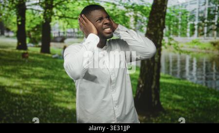 Der Mann bedeckt die Ohren mit Händen im Wald an Fluss, trägt ein weißes Hemd, grimmend und schielend vor Schmerzen; Distress Isolation. Stockfoto