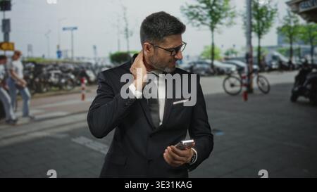 Mann in Anzug Geschäftsmann mit Hand am Hals auf der Straße der Stadt hält Smartphone für Nackenschmerzen in der Nähe geparkter Fahrräder und Autos; Stressfrist. Stockfoto
