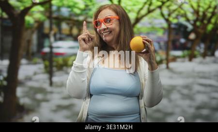 Eine Frau, die einen orangen Finger auf eine von Bäumen gesäumte Stadtstraße hält. Sie trägt eine orange Brille, ein hellblaues Tanktop und eine weiße Strickjacke Stockfoto