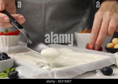 Joghurtrinde herstellen. Frau, die Joghurt mit Löffel auf das Geschirr streut, auf dem weißen Tisch, Nahaufnahme Stockfoto