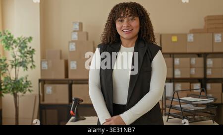 Die hispanische Frau lächelt mit einem Pappkarton neben einem Barcode-Scanner und einem Regal in einem Bürogebäude; Vertrauen in Unternehmertum. Stockfoto
