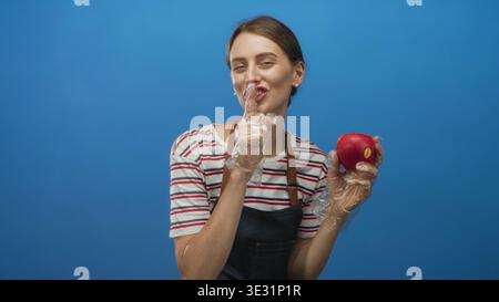 Frau in Schürze und Einweghandschuhen, die roten Apfel und Finger im blauen Atelier an die Lippen hält; verspielte Geheimhygiene. Stockfoto
