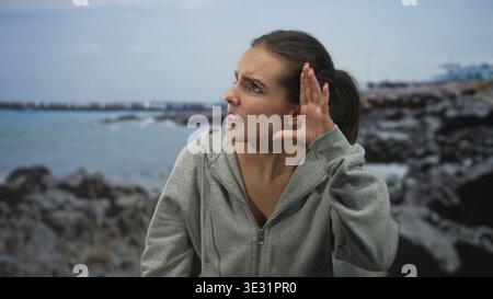 Junge hispanische Frau, die mit der Hand das Ohr schröpft und im Studio am Meer schielt, mit grauem Kapuzenpullover; frustrierte Hörschwierigkeiten. Stockfoto