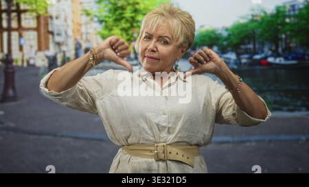 Frau Senior zeigt Daumen nach unten, beide Hände nahe dem Gesicht und sichtbare Armbänder auf einer Kopfsteinpflasterstraße an einem Kanal; Missbilligung. Stockfoto