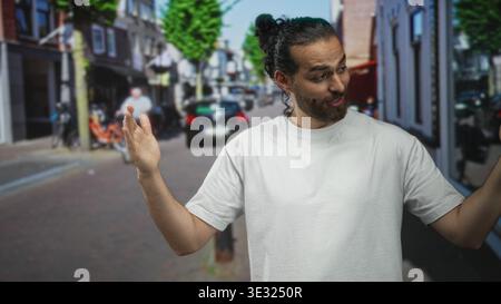 Mann, der mit geöffneten Palmen und erhobenen Händen auf einer sonnigen Stadtstraße mit weißem T-Shirt und Brötchen-Frisuren gestickt hat; verspieltes Zucken. Stockfoto