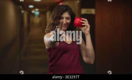 Die junge hispanische Frau zeigt mit dem Finger nach vorne, während sie in einem Hotelgebäude eine rote Schweinebank hält. Stockfoto