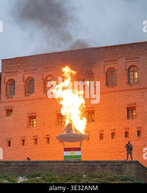 Erbil, Region Kurdistan, Irak. März 2026. Flammen und dicker Rauch steigen aus einer zeremoniellen Fackel auf, die in den Farben der kurdischen Flagge während der Newroz-Feiern vor einer historischen Zitadelle verziert ist Sicherheitspersonal halten Wache in der Nähe des massiven Lagerfeuers, das zum Gedenken an das kurdische Neujahr und die Ankunft des Frühlings angezündet wird (Credit Image: © Mahmood Yaseen/ZUMA Press Wire). Nicht für kommerzielle ZWECKE! Stockfoto