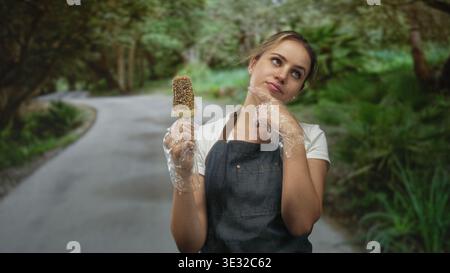 Frau mit Eis und Schürze mit Einweghandschuhen, Finger an Wange auf einer Waldstraße; Versuchungszweifel. Stockfoto