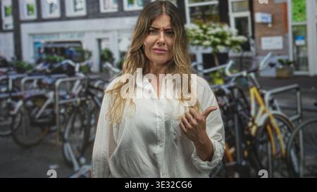 Die Frau zeigt mit dem Daumen auf geparkte Fahrräder auf der Stadtstraße, während sie die Stirn runzelt und unzufrieden aussieht; Frustration. Stockfoto