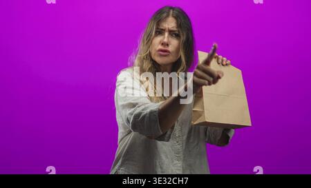 Frau zeigt mit dem Finger und hält die braune Papiertüte vor violettem Studio-Hintergrund, während sie nach links drückt; Frustration. Stockfoto