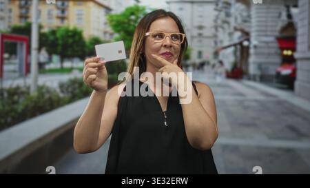 Frau mit weißer Kreditkarte zeigt Finger zum Kinn auf der geschäftigen Straße der Stadt; durchdachte Kaufentscheidung. Stockfoto