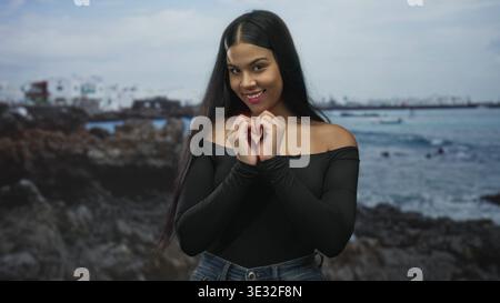 Frau, die Herz mit Händen an der felsigen Küste unter bewölktem Meereshimmel bildet; Liebe Liebe Liebe Freude Romantik. Stockfoto