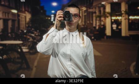 Die Frau hält die dslr-Kamera in der nächtlichen Straße, trägt ein weißes Hemd und eine rote Brille, eingerahmt von geparkten Fahrrädern und Café-Lichtern; kreativer Fokus. Stockfoto