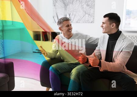 Glückliches Schwulenpaar, das sich auf dem Sofa zu Hause unterhalten hat, doppelte Exposition mit Stolz-Flagge Stockfoto