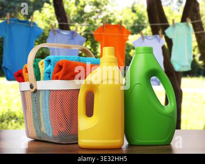 Korb mit Wäsche und Flaschen Waschmittel auf Holztisch in der Nähe von Kleidung, die draußen trocknet Stockfoto