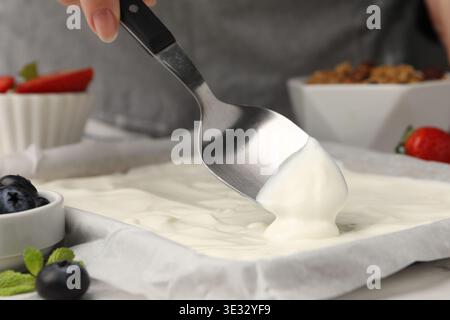 Joghurtrinde herstellen. Frau, die Joghurt mit Löffel auf das Geschirr streut, auf dem weißen Tisch, Nahaufnahme Stockfoto