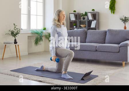 Ältere Frau, die während des Online-Trainings Sport mit Kurzhanteln auf Yogamatte mit Tablet macht. Stockfoto