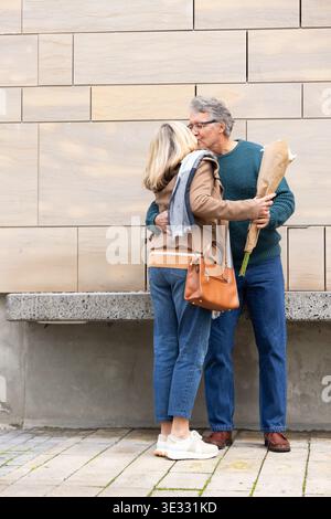 Seniorenpaar umarmen, küssen vor der beigefarbenen Wand, Mann mit Blumenstrauß, Frau mit Handtasche Stockfoto