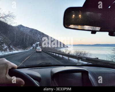 Blick aus dem Auto mit einer Hand am Lenkrad während der Fahrt auf einer Landstraße in der Nähe des Baikalsees in der Abenddämmerung. Stockfoto