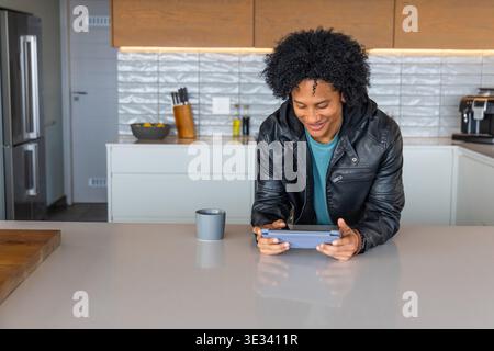 Afroamerikanischer Erwachsener, der sich auf der Kücheninsel mit Tablette stützt und eine schwarze Lederjacke trägt Stockfoto