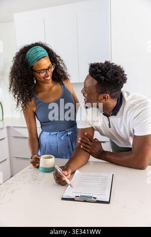 Afroamerikanisches Paar steht auf der Kücheninsel, lächelnd, hält einen zweifarbigen Becher und ein Klemmbrett Stockfoto