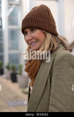 Ältere Frau, die auf der Dachterrasse nach rechts steht, trägt braune Beanie, Schal, Olivenmantel Stockfoto